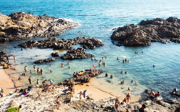 Vibrant beach scene with people enjoying a rocky shore cove under sunny skies.