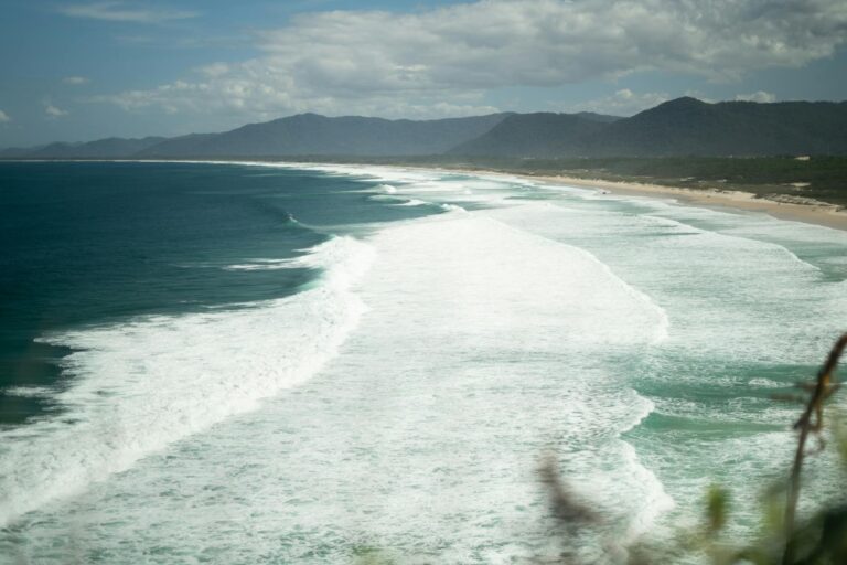 A scenic view of Florianópolis beach with waves crashing against the shore and mountains in the background.