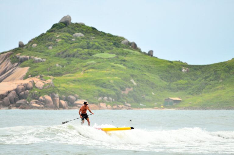 A man paddleboarding on waves in Barra Da Lagoa, Brazil, with lush green hills in the background.