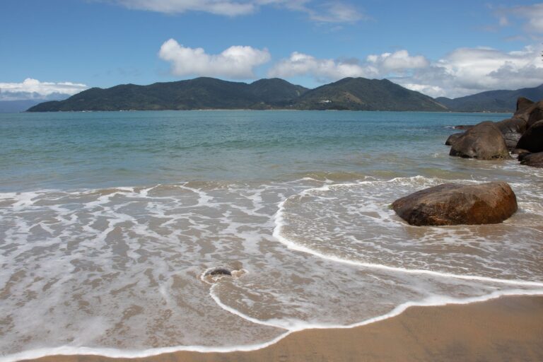 Tranquil beach with boulders and distant mountains under a clear blue sky.