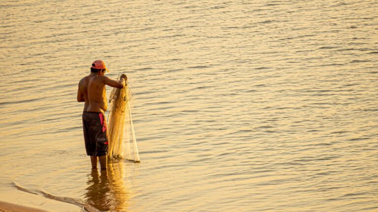 A fisherman casts his net at sunset on the serene waters of Pindobal, Brazil.