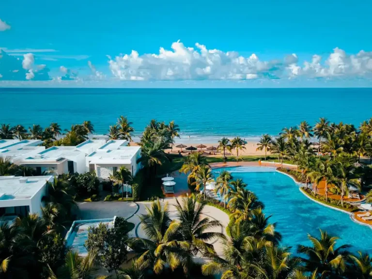 Aerial view of a luxurious beachfront resort with palm trees, pool, and ocean view.